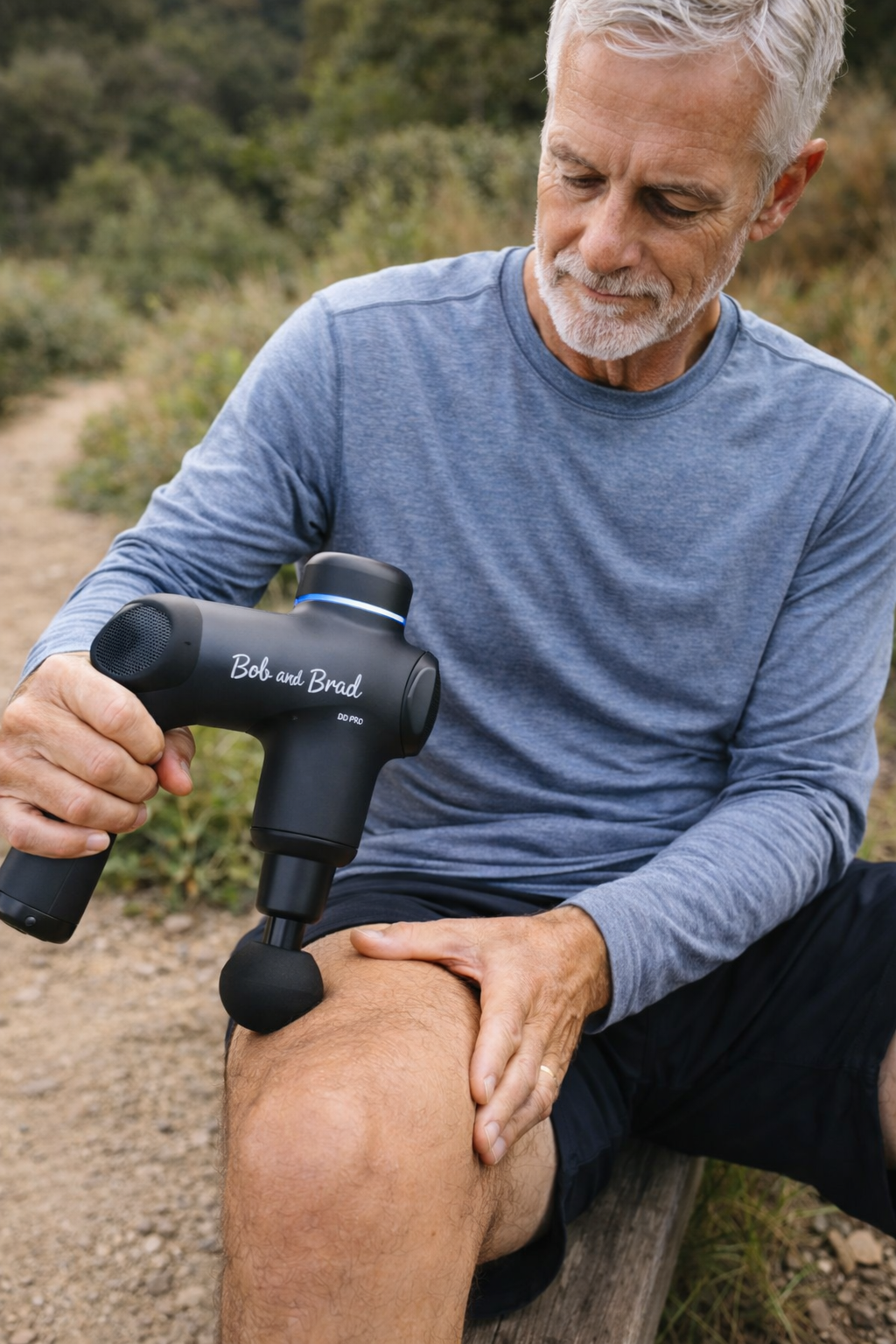 Older man using a massage gun on his knee while sitting on a bench on a trail
