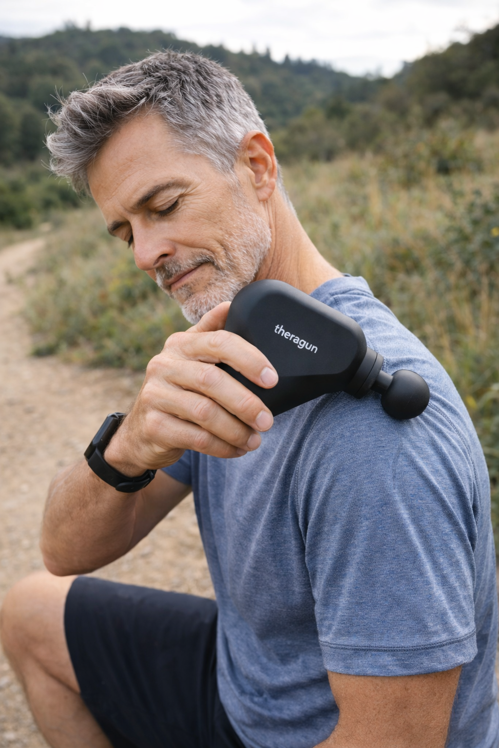 Man using a massage gun on his shoulder while sitting on a trail