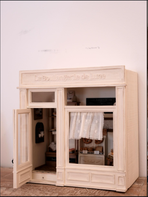 Pale miniature bakery storefront with an open door and curtained window