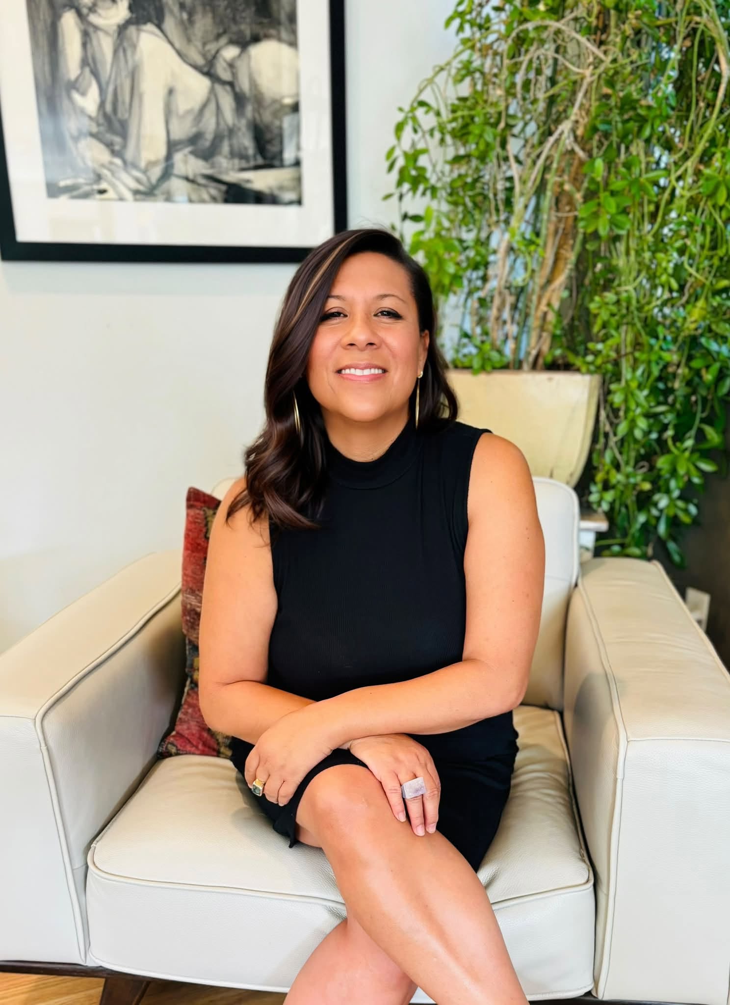 Janice Noehulani seated in a light armchair beside a leafy plant