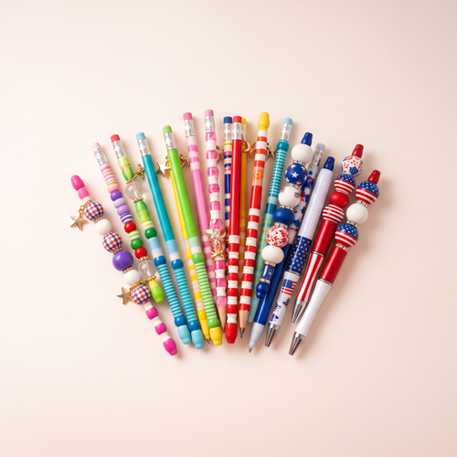 Colorful assortment of beaded pens and pencils with decorative beads and charms on a warm ivory and blush-pink boutique background