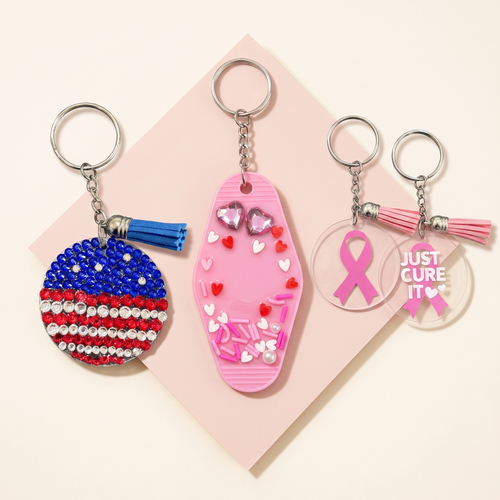Three keychains: a blue, red, and white beaded keychain, a pink heart keychain, and two pink ribbon keychains, one reading JUST CURE IT, on a warm ivory and blush background