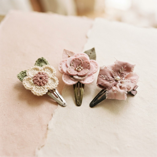 Three handmade flower hair clips in pastel pink, warm ivory, and dusty rose with visible metal clips on a blush and ivory background