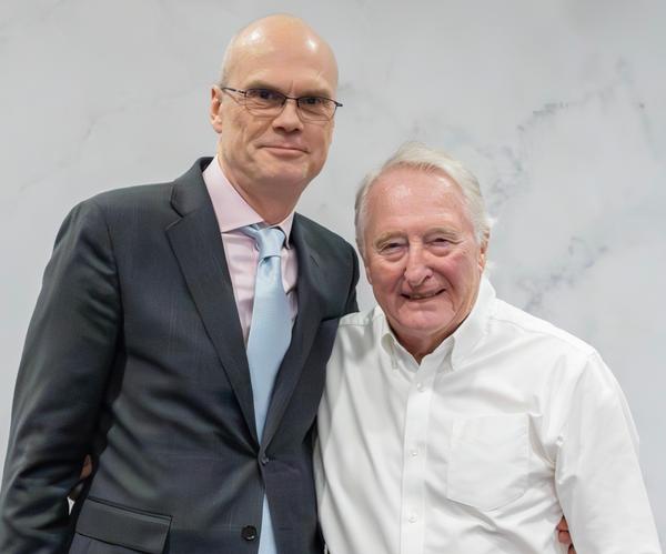 Two men posing together, one in a dark suit and one in a white shirt