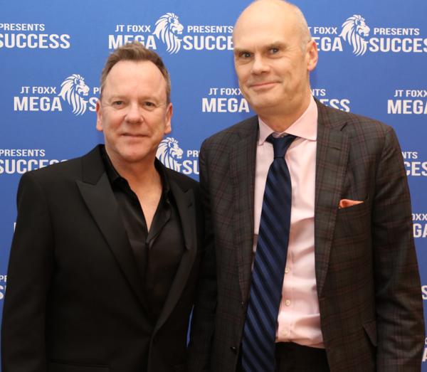 Two men in formalwear posing before a blue event backdrop