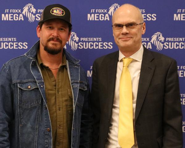 Two men posing together in front of a blue event backdrop, one wearing a cap