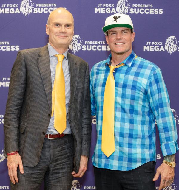 Two men posing together in front of a purple event backdrop, one wearing a checked shirt