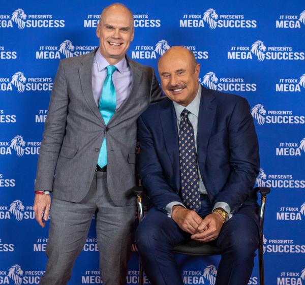 Two men posing together in front of a blue event backdrop, one seated