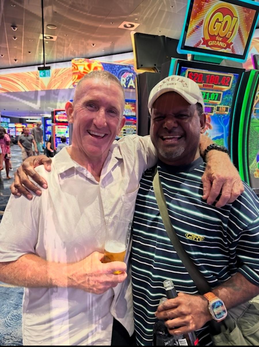 Misi smiling beside another man in a striped shirt in a brightly lit indoor venue with gaming machines in the background.